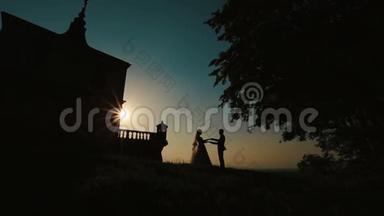 在古堡附近的夕阳下，幸福新婚夫妇温柔地牵手亲吻的剪影。